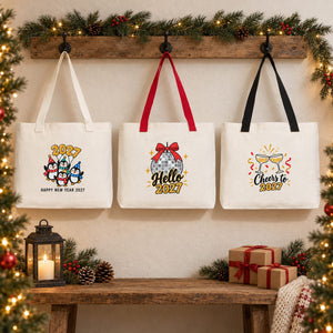 Three Christmas 2027 canvas tote bags with beige, red and black knitted straps hanging on a rustic wall with penguin, Hello 2027 disco ball and Cheers to 2027 prints
