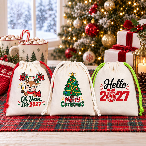Three natural cotton drawstring gift pouches with red, natural and green cords, printed with Oh Deer It's 2027, Merry Christmas tree and Hello 2027 designs, on a tartan table with hot cocoa, a knitted stocking and a Christmas tree