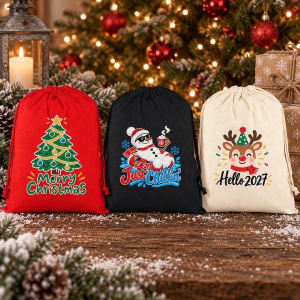 Three Christmas drawstring gift bags in red, black and natural cotton with gathered tops and hanging cords, printed with Merry Christmas tree, 2027 Just Chillin snowman and Hello 2027 reindeer designs, on a snowy rustic table in front of a lit Christmas tree