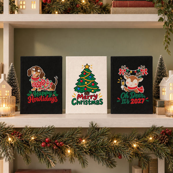 Three A5 canvas notebooks, black and natural, with Happy Howlidays dachshund, Merry Christmas tree and Oh Deer It's 2027 reindeer prints on a Christmas bookshelf