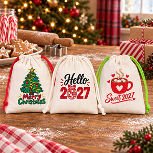 Three 4 x 5 inch cotton drawstring favour bags with red, natural and green cords printed with Merry Christmas, Hello 2027 and Sweet 2027 designs on a flour-dusted kitchen table with gingerbread cookies and candy canes