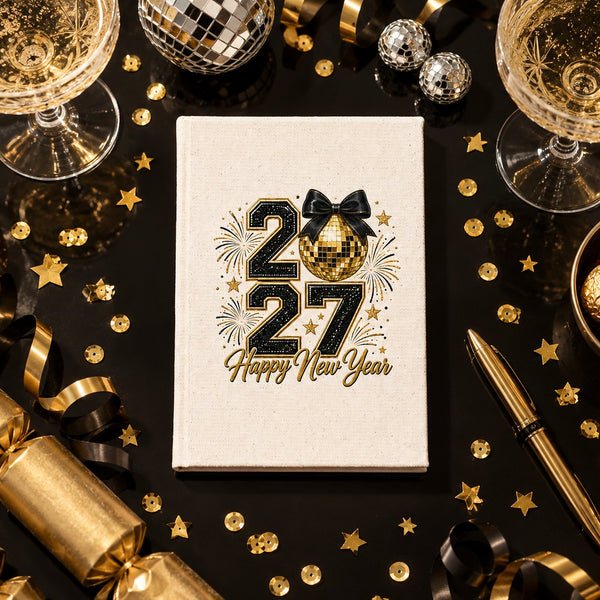 Natural canvas notebook with a 20/27 Happy New Year gold disco ball and bow print on a black New Year's Eve party table with champagne and gold confetti