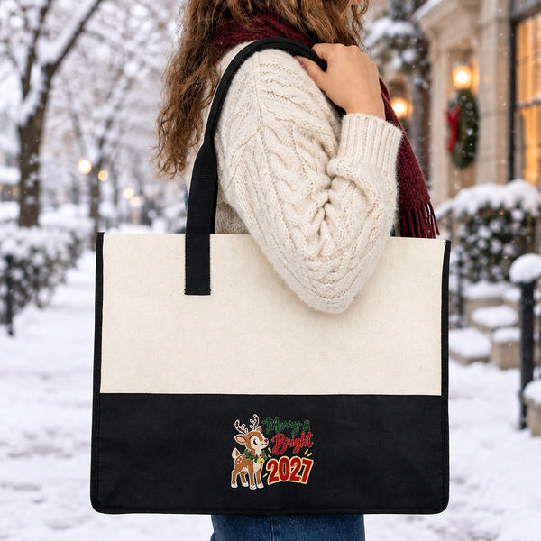 Woman in a cream cable-knit wool sweater and burgundy scarf carrying the black canvas tote bag with a Merry and Bright 2027 reindeer print on her shoulder on a snowy winter street
