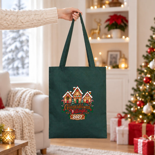 Hand lifting a forest green cotton tote bag with a Gingerbread Village 2027 print in a bright Christmas living room