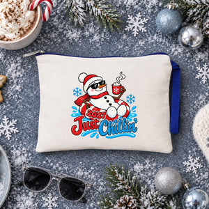 Cream zipper pouch with a royal blue zipper and a Just Chillin 2027 snowman print on a snowy blue-grey surface with cocoa and mittens