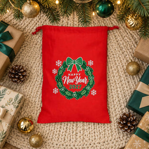 Red drawstring gift bag with a Happy New Year 2027 holly wreath print lying on a cream knit tree skirt among wrapped presents under the Christmas tree