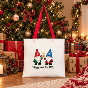 Cotton tote bag with red handles and a Happy Gnome Year 2027 print standing under a Christmas tree among wrapped gifts