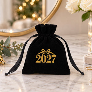 Black velvet jewellery pouch printed with a gold bow 2027 design on a marble vanity beside a ring box, gold rings and a pearl necklace