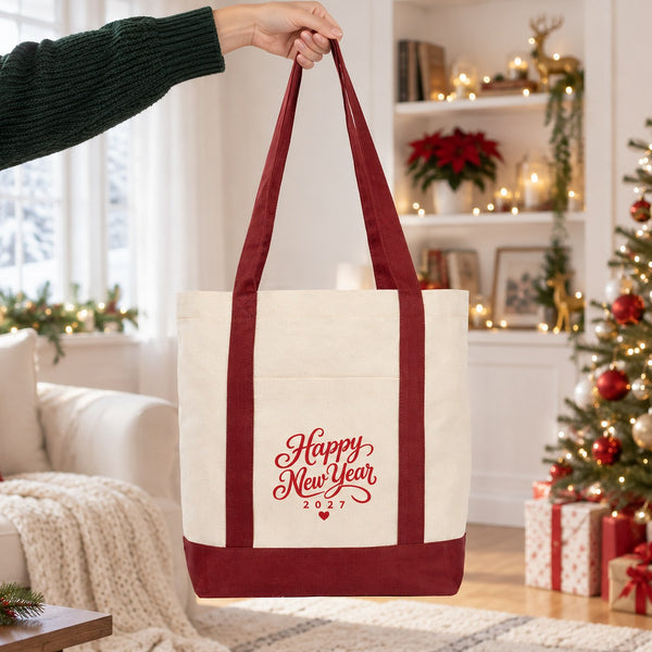 Hand lifting a burgundy strap canvas tote bag with a red Happy New Year 2027 script print in a bright Christmas living room