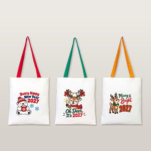 Three natural cotton tote bags with red, green and mustard handles and Beary Happy New Year, Oh Deer and Merry and Bright 2027 prints on a plain studio background
