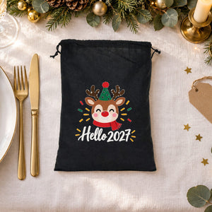Black drawstring bag with a Hello 2027 reindeer print lying beside a place setting on a Christmas dinner table as a guest party favor