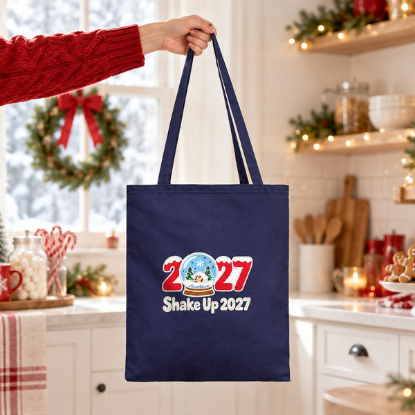 Hand lifting a navy blue cotton tote bag with a Shake Up 2027 snow globe print in a bright Christmas kitchen