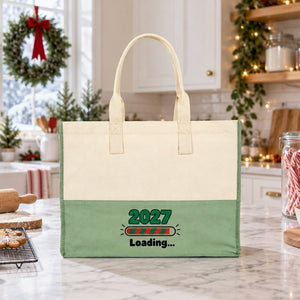 Sage green canvas tote bag with a 2027 Loading print standing on a marble kitchen counter with gingerbread cookies and candy canes