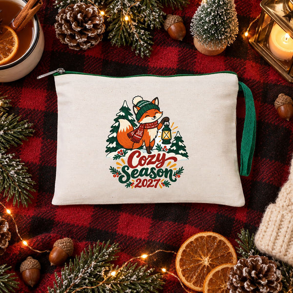 Cream clutch with a green zipper and a Cozy Season 2027 fox print on a buffalo plaid blanket with a lantern, pine cones and cider
