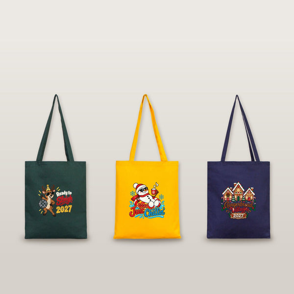 Studio photo of three cotton gabardine tote bags in green, yellow and navy with Ready to Sleigh 2027, Just Chillin' 2027 and Gingerbread Village 2027 prints