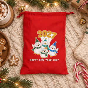 Red drawstring bag with a Happy New Year 2027 penguins print on a cream knit blanket with hot cocoa, gingerbread cookies, candy canes and a knit stocking