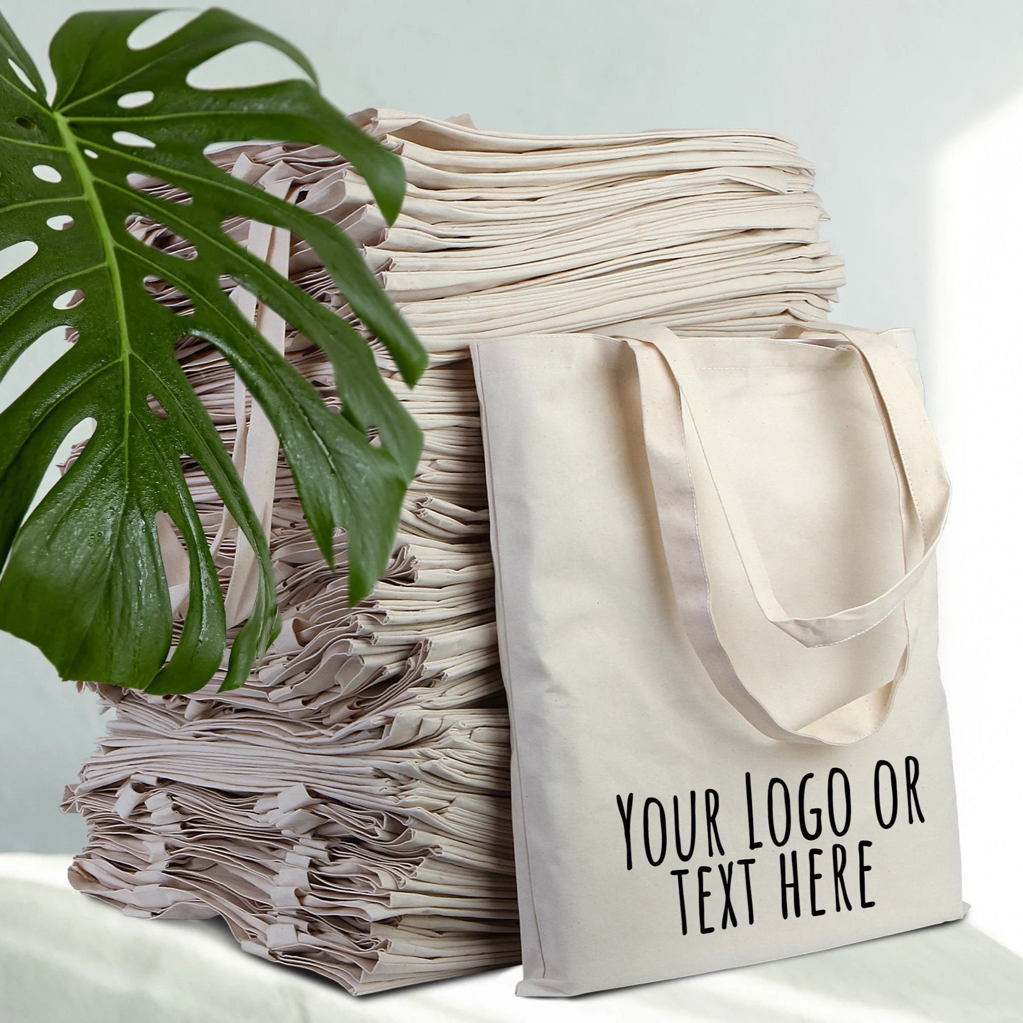 Beige tote bag with customizable text or logo on a white surface with green leaves.