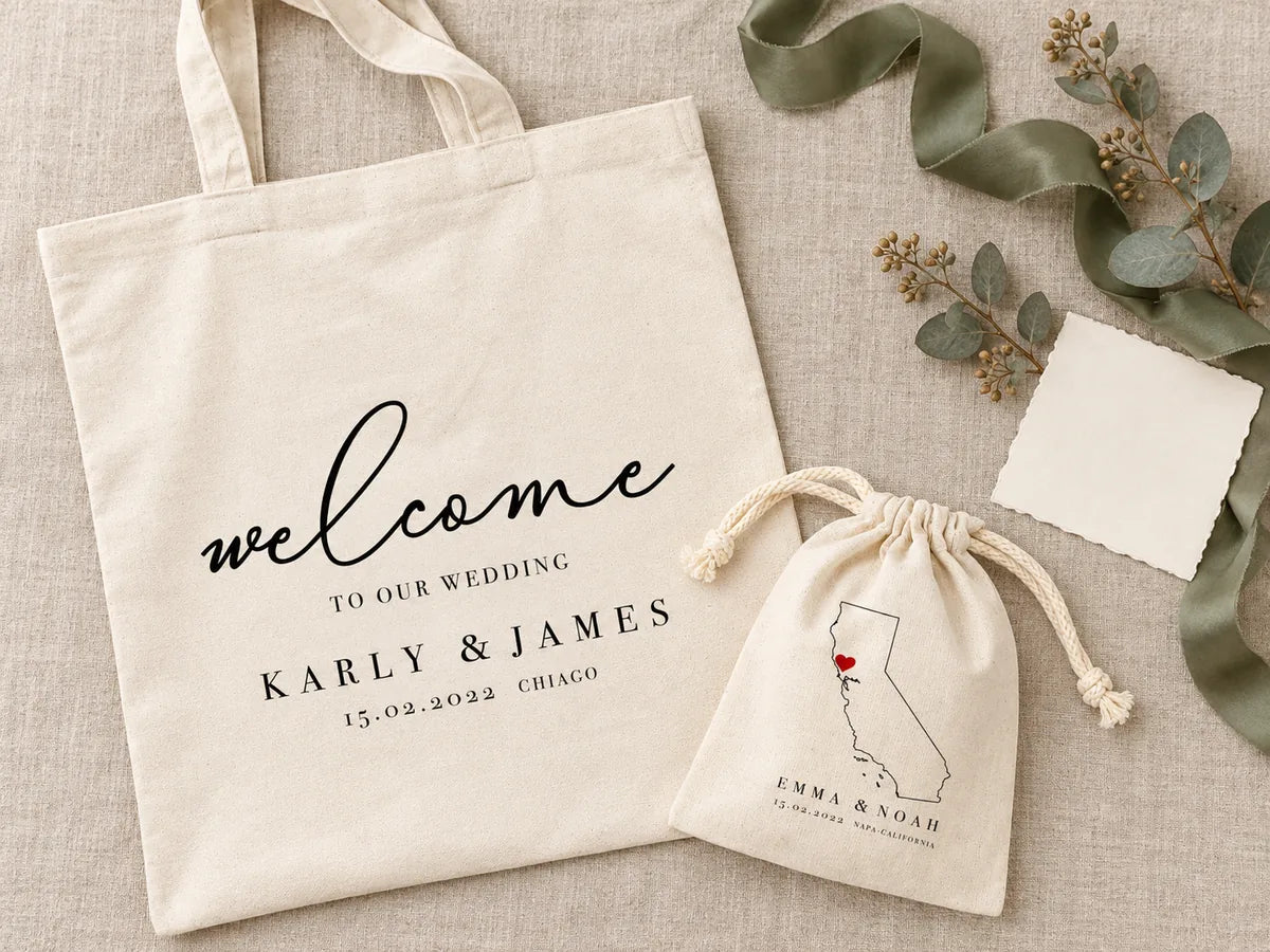 Flat-lay of a welcome tote printed with names and date beside a favor pouch, sage ribbon and eucalyptus on linen